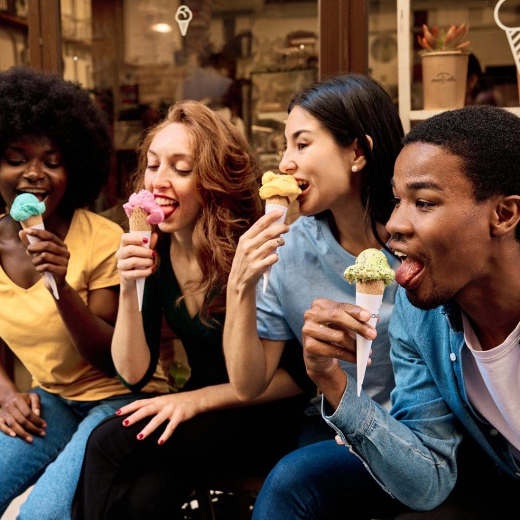 Four people sitting at a bench reaching their tongues out to lick ice cream cones.